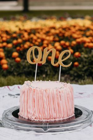 A pink ruffle cake with gold 'one' cake topper surrounded by orange flowers.