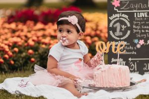 Baby wearing pink tutu and headband sitting next to pink birthday cake outdoors.
