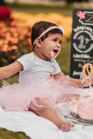 Baby in white outfit and pink tutu enjoying birthday cake smash outdoors.