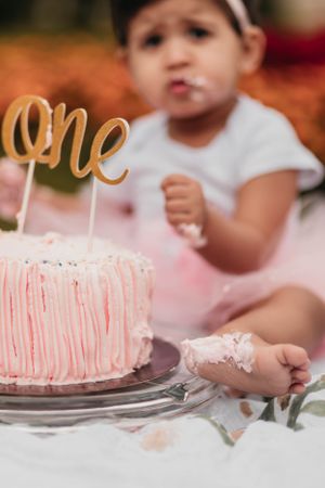 Close-up of a pink birthday cake with gold 'one' topper and small hand reaching for it.