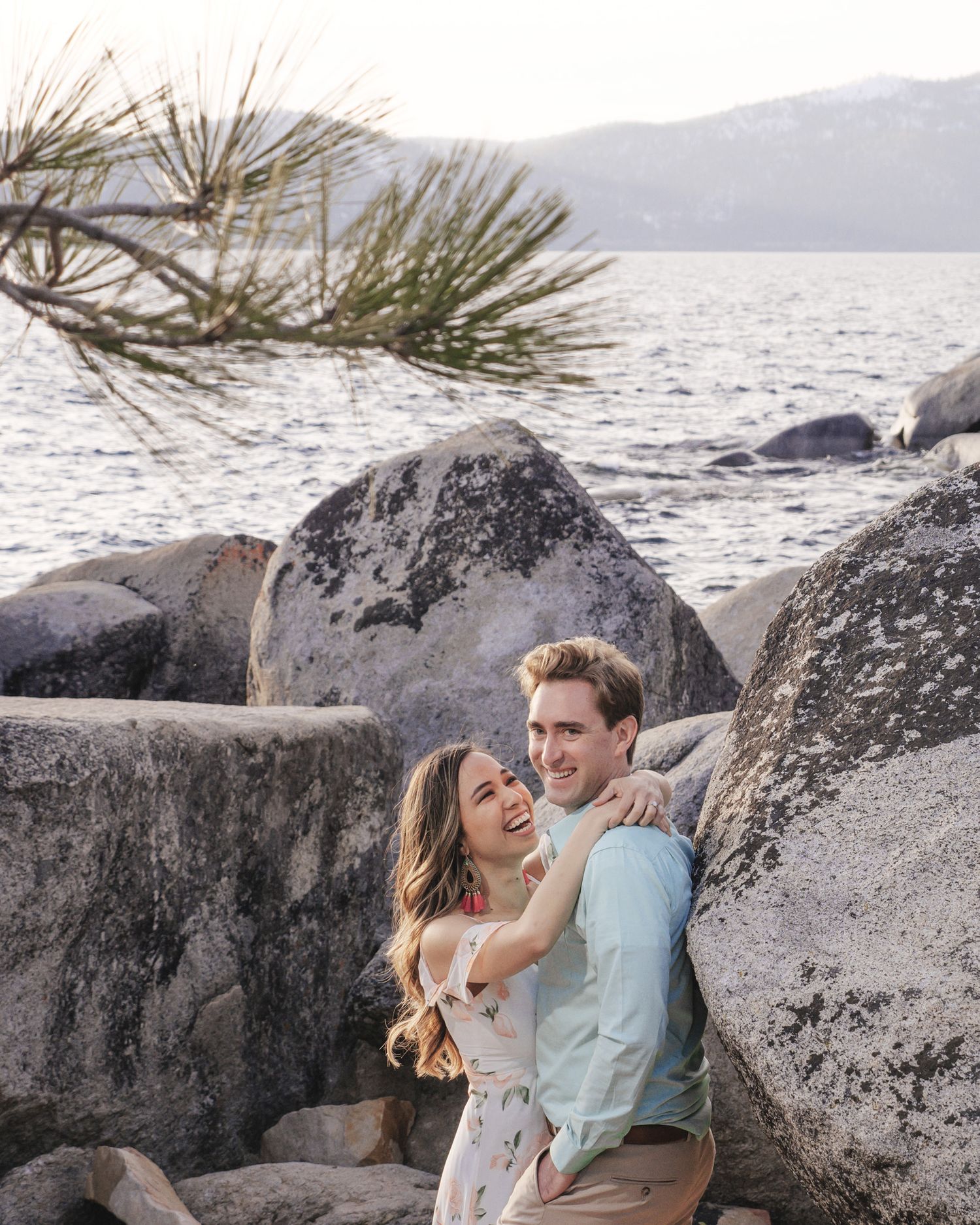 A couple embraces amongst large boulders along a serene Lake Tahoe shore with pine needles and mountains in the background.