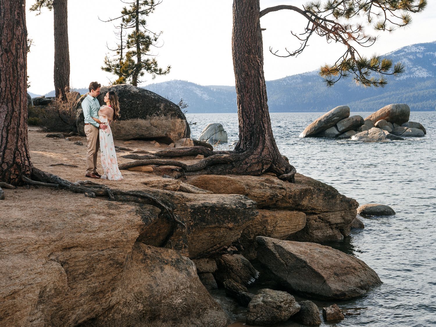 A couple shares a romantic moment on rocky cliffs overlooking Lake Tahoe surrounded by towering pine trees at sunset.