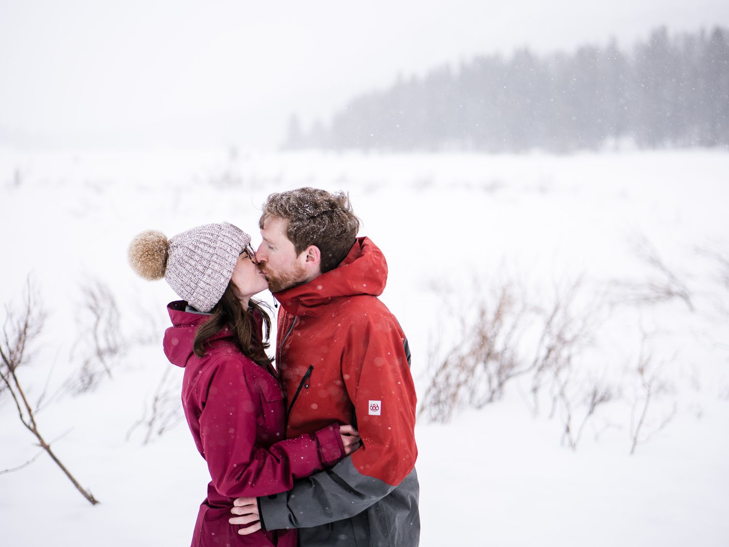 A couple sharing a romantic moment in winter, wearing red and burgundy coats against a snowy landscape background.