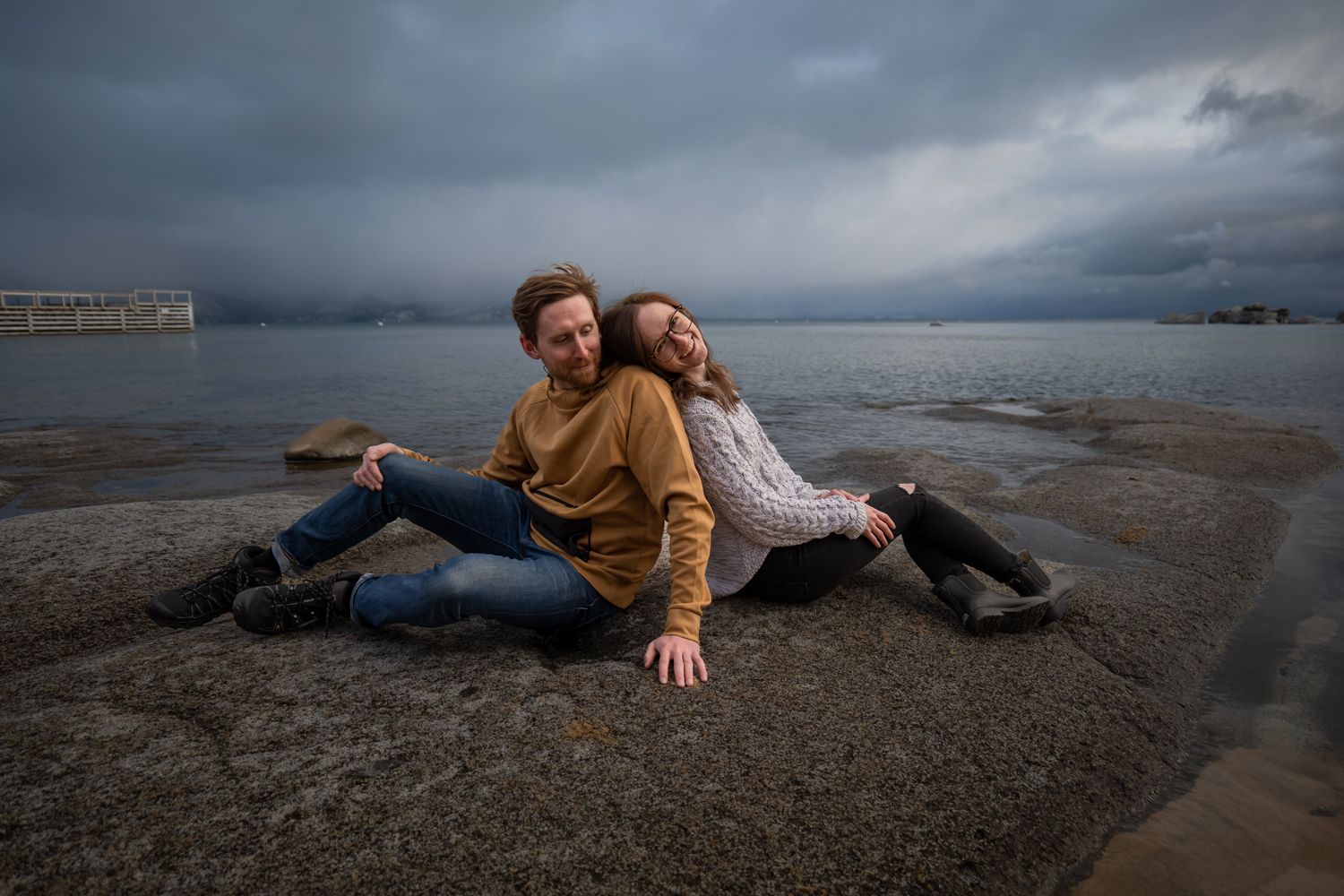 Couple sitting together on rocky beach against dramatic cloudy sky and waterfront during moody evening.