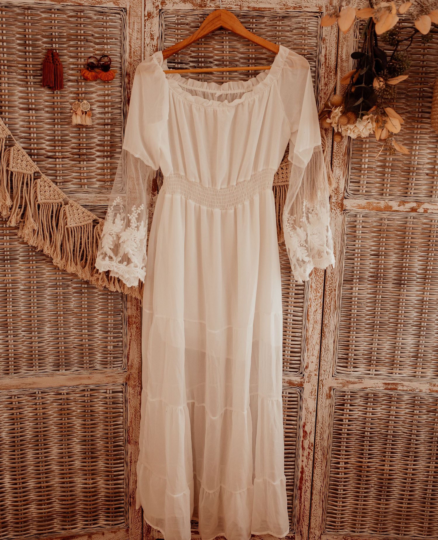 White cotton dresses with feminine details displayed on wooden hangers against wicker wall.