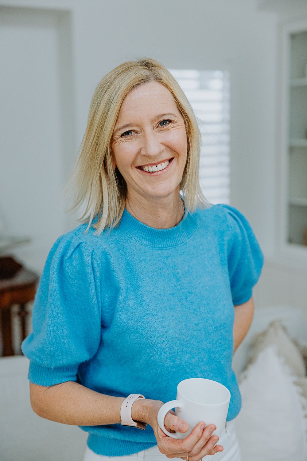 Casual portrait of someone in a bright blue sweater holding a white coffee mug in a bright interior space.