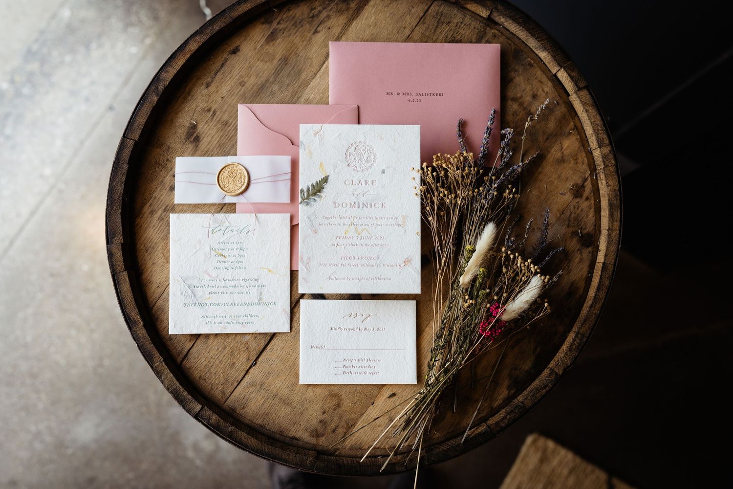 Wedding invitation suite with pink stationery and dried flowers displayed on a rustic wooden barrel.