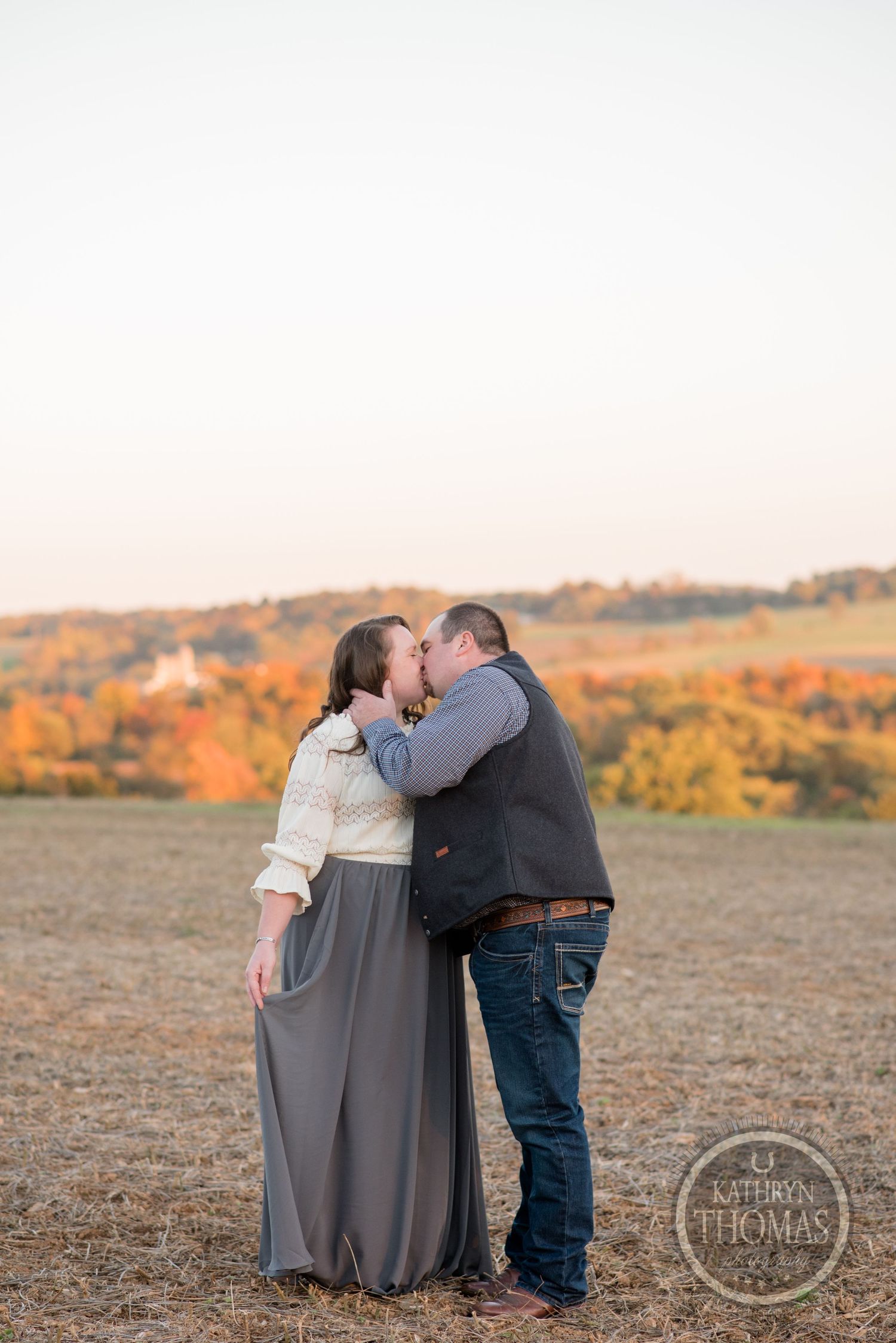 Couple sharing a romantic embrace in a field during golden hour with autumn foliage in background.