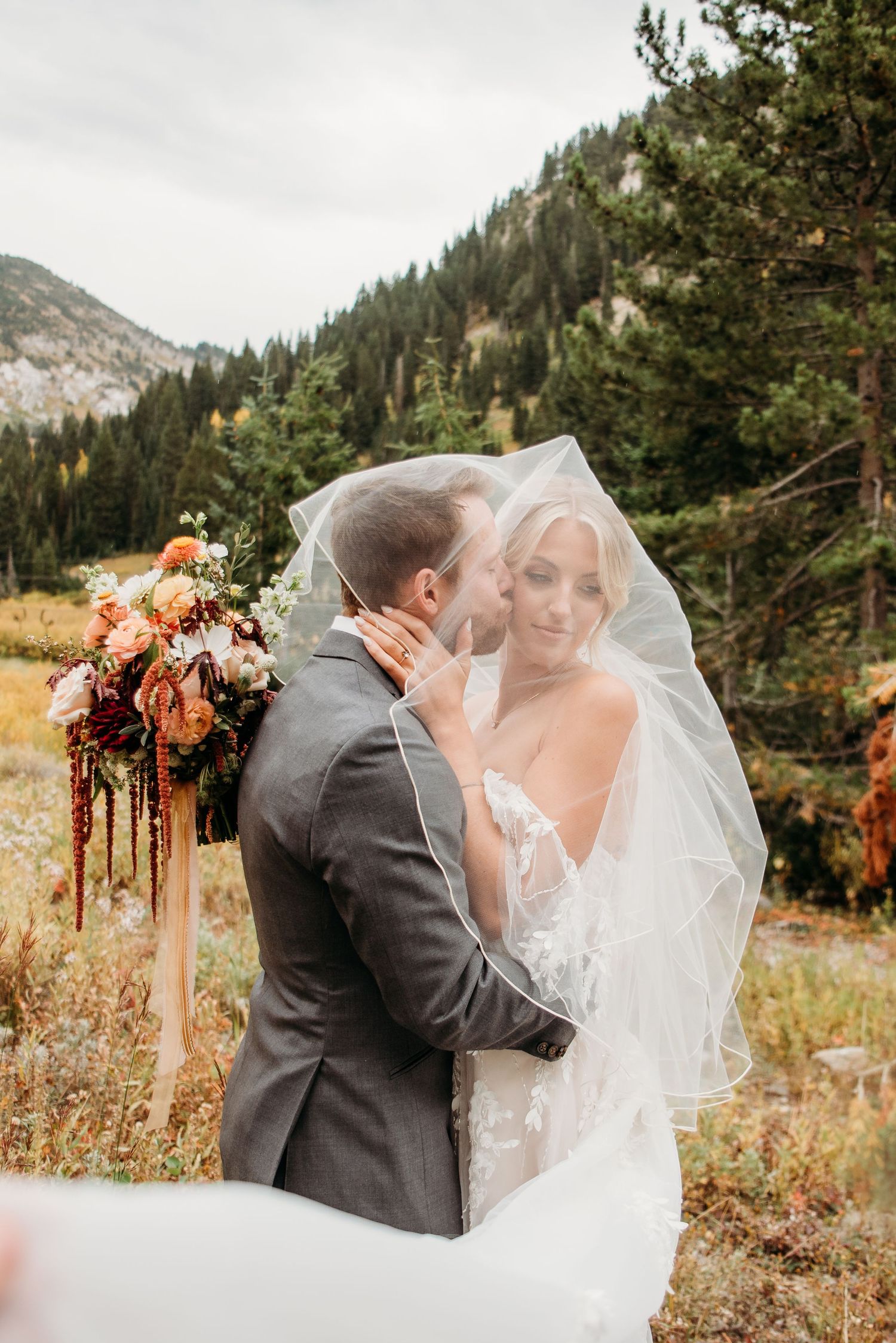 A romantic wedding moment captured in a mountainous forest setting with warm autumn colors and flowing veil.
