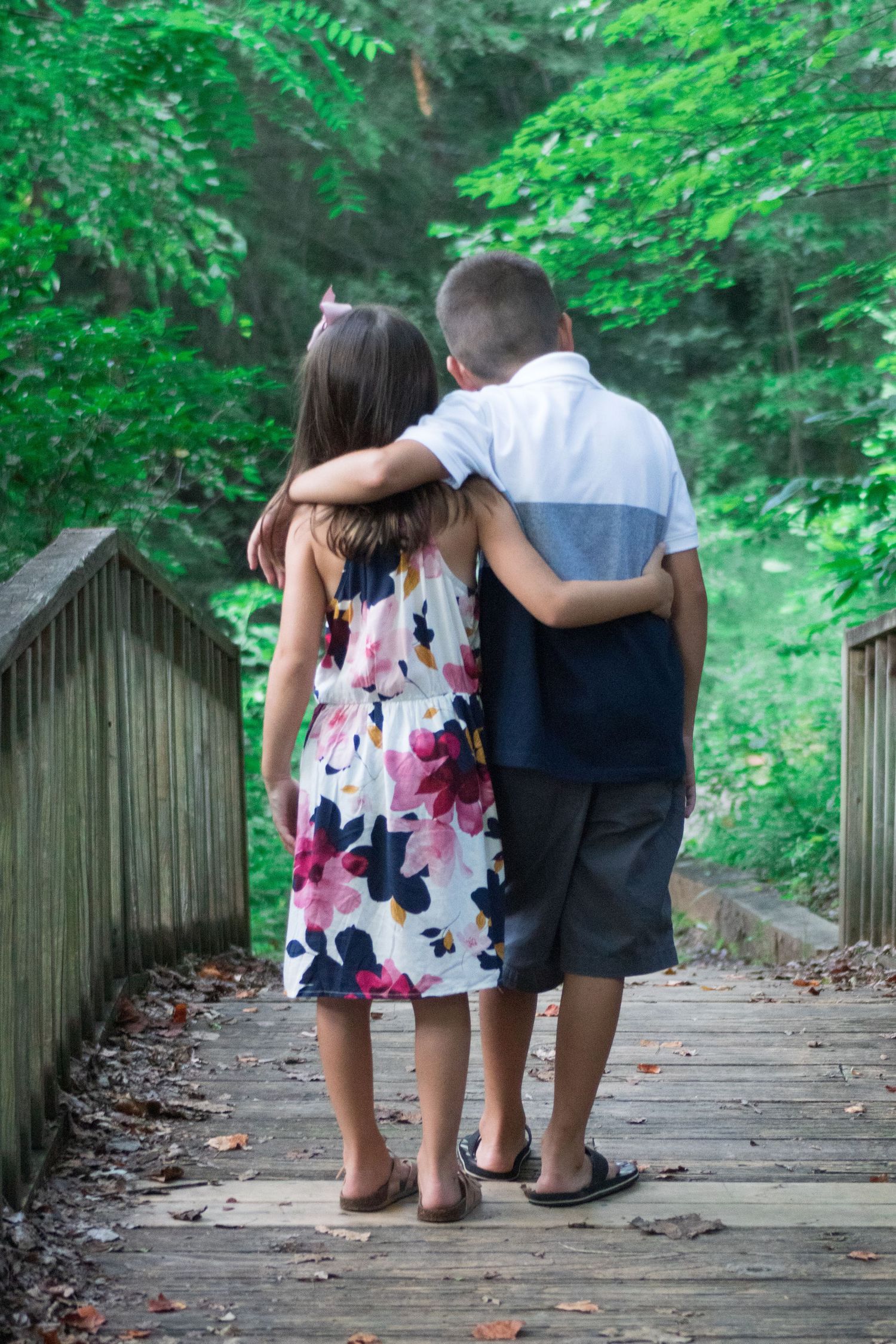 Two people walk together on a wooden bridge path through lush green forest.