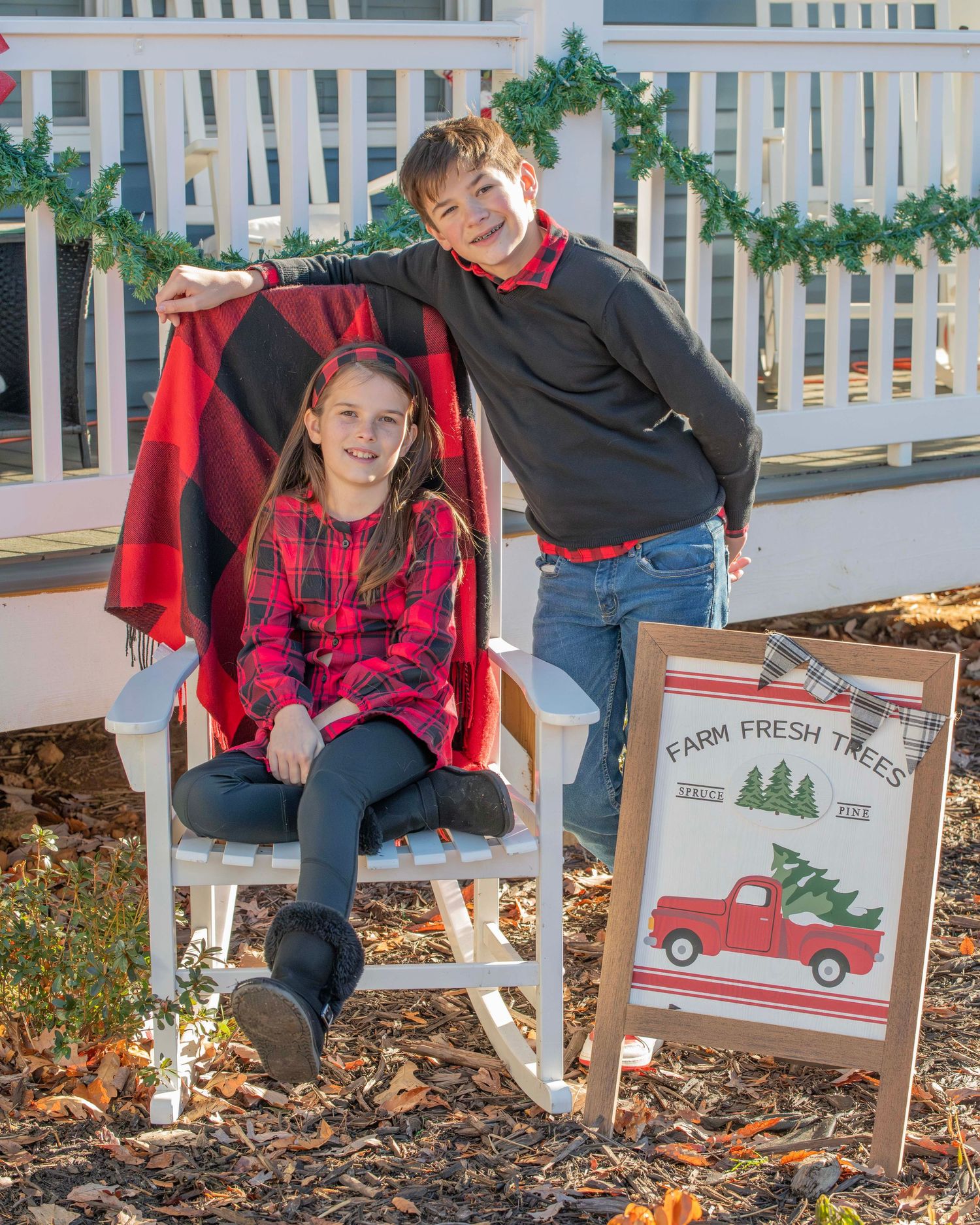 Holiday portrait with two people by a decorated porch with a Christmas tree farm sign.