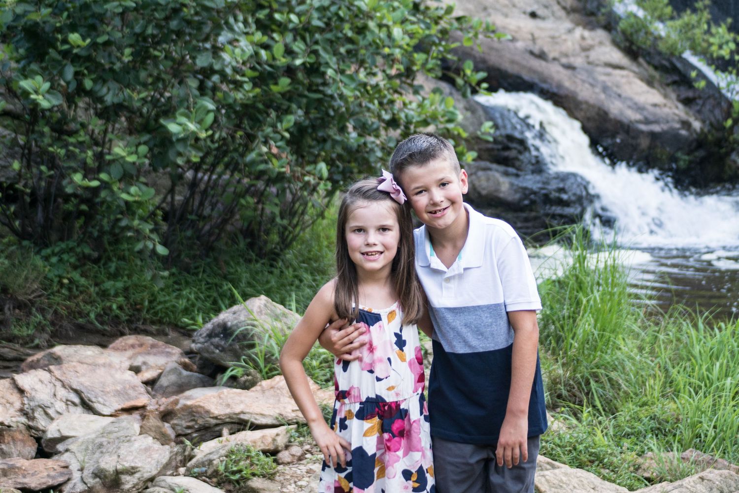 Two young people pose in front of a cascading waterfall on a scenic forest trail.