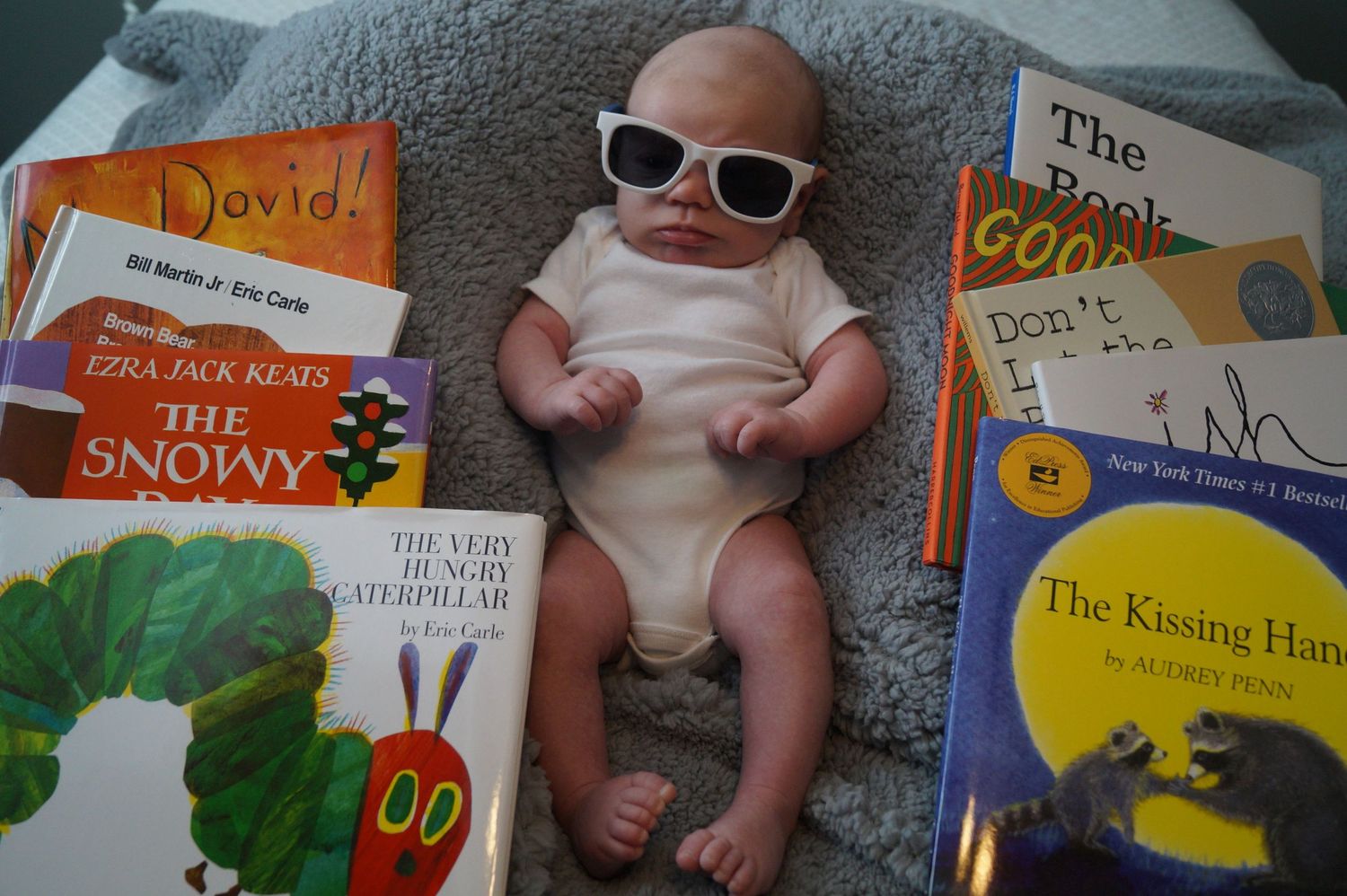 Baby lying on gray blanket surrounded by colorful children's books showing storybook covers and spines.