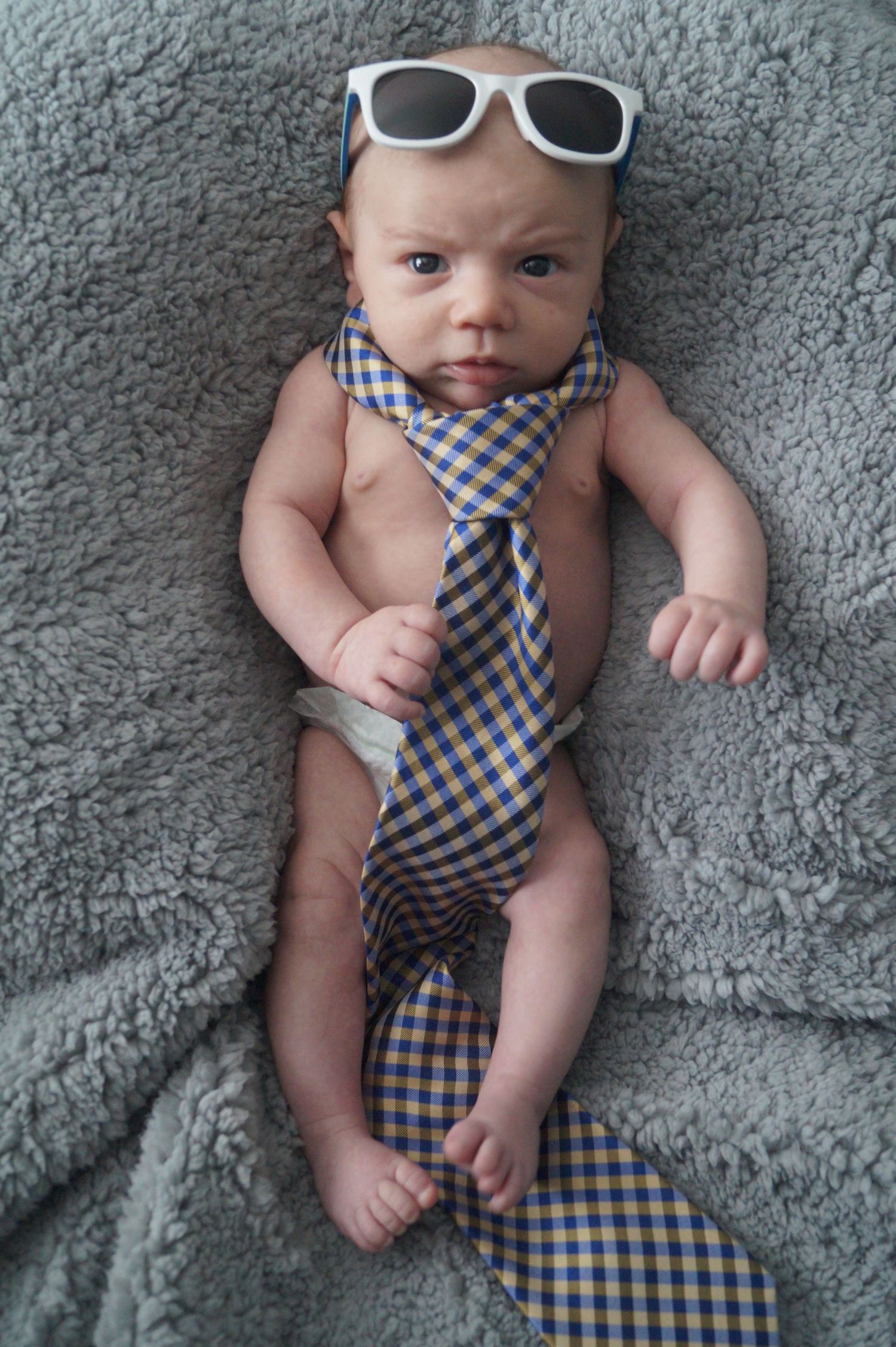 Baby wearing checkered tie and sunglasses while lying on plush gray carpet.