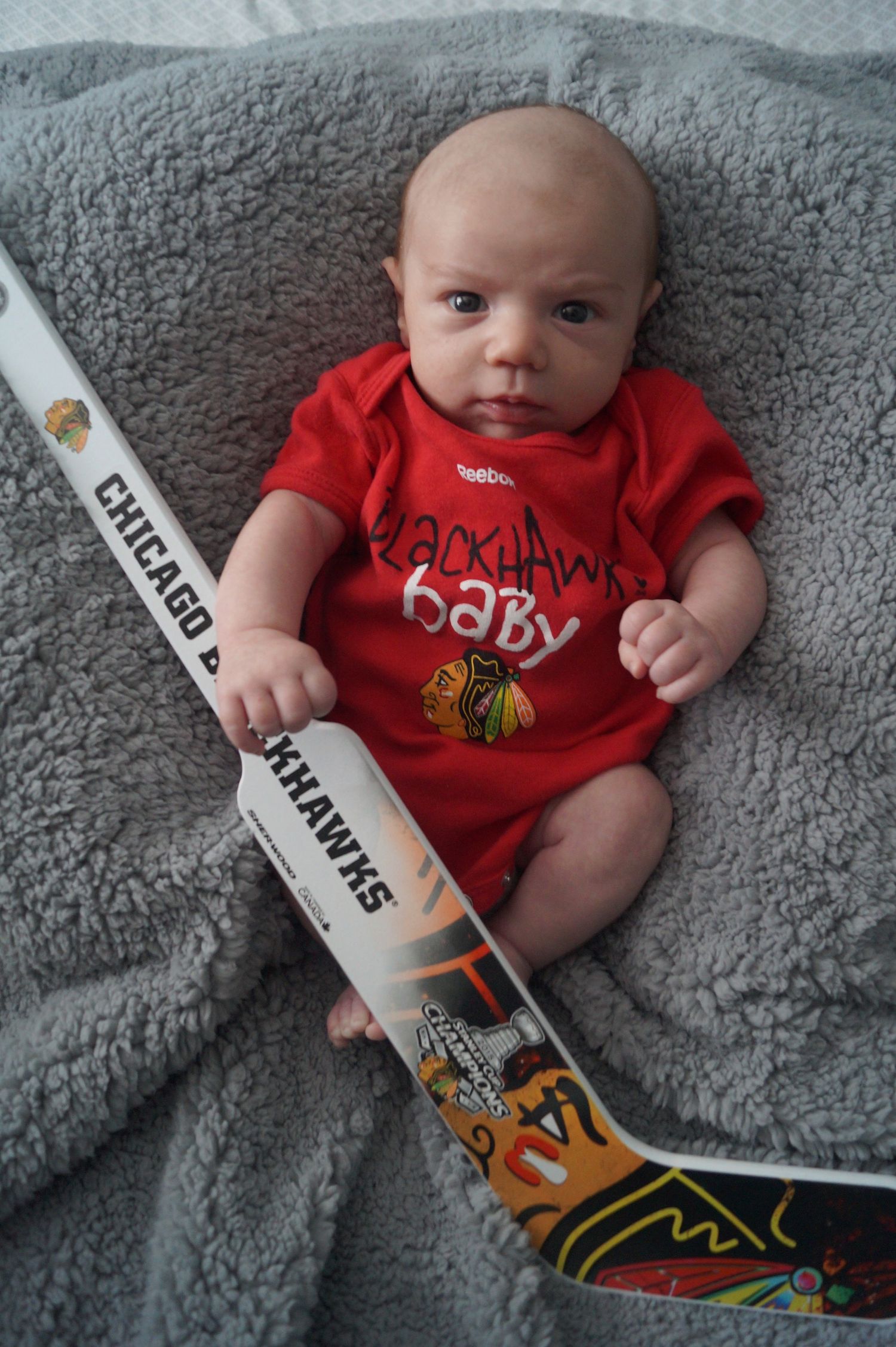 Baby wearing red sports jersey with hockey sticks placed nearby on gray background.