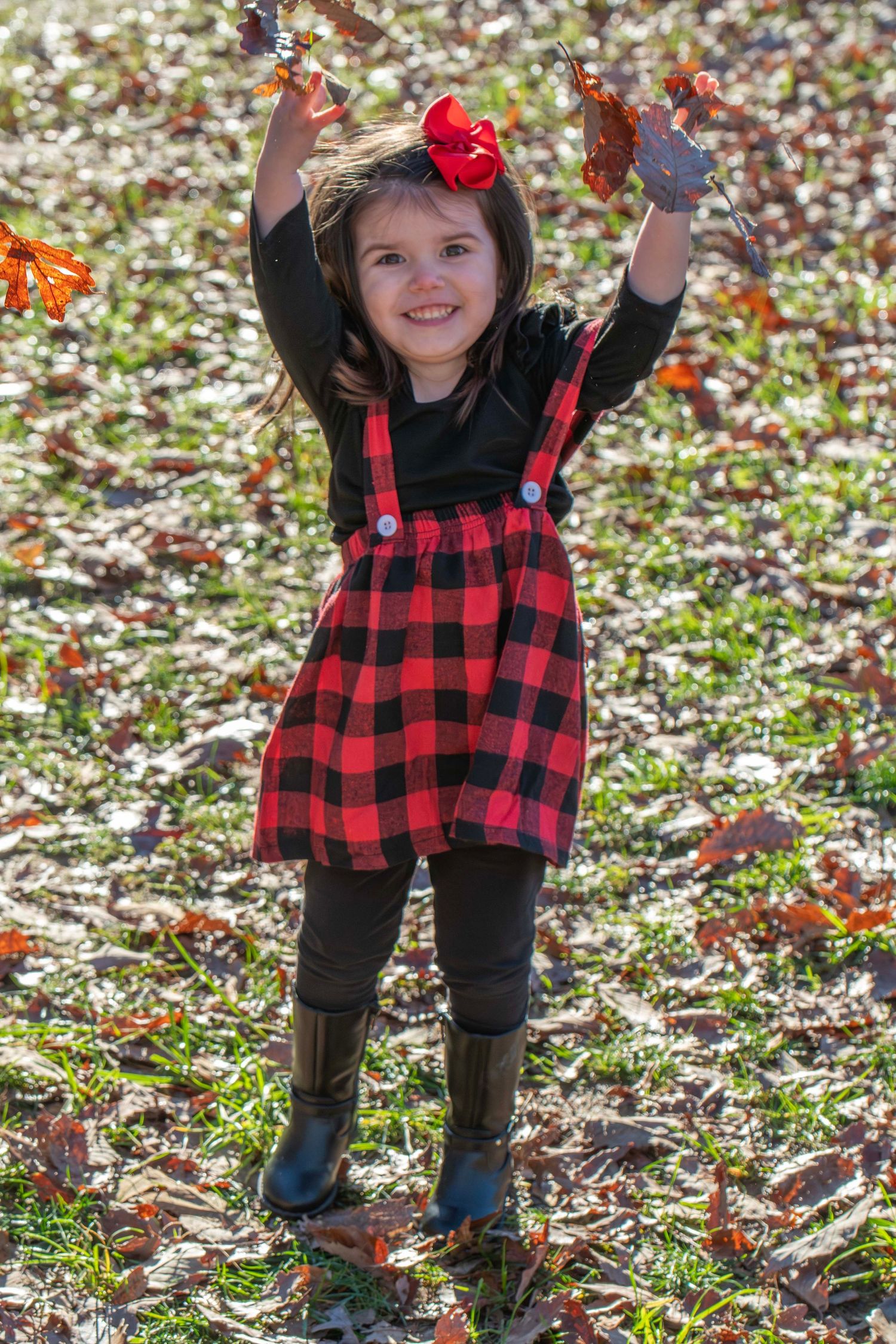 Young person in buffalo plaid dress throws autumn leaves while playing outdoors.