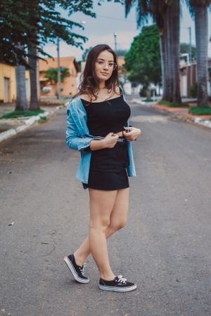 Street fashion photoshoot showcasing black top and denim jacket with sneakers on urban sidewalk.