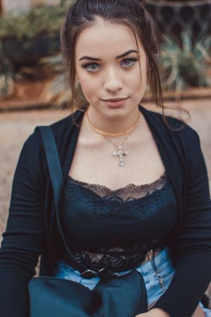 Close-up portrait showing lace top, black cardigan and cross necklace against garden background.
