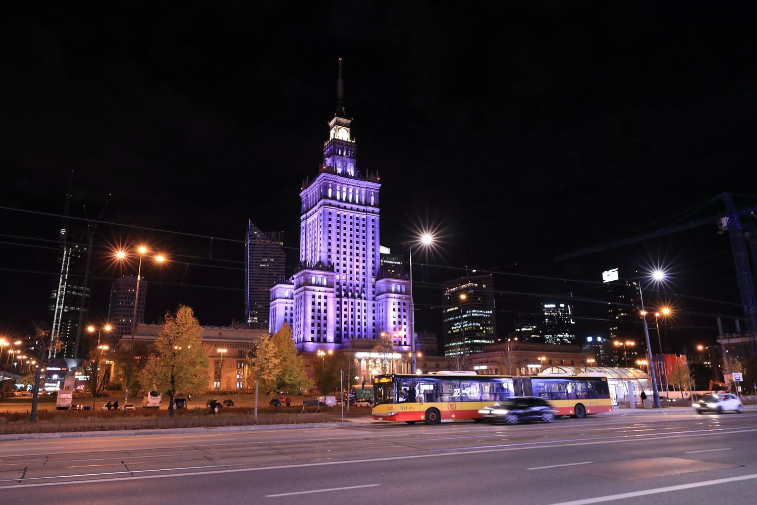 Palace of Culture and Science illuminated in purple light at night with traffic and trams in Warsaw.