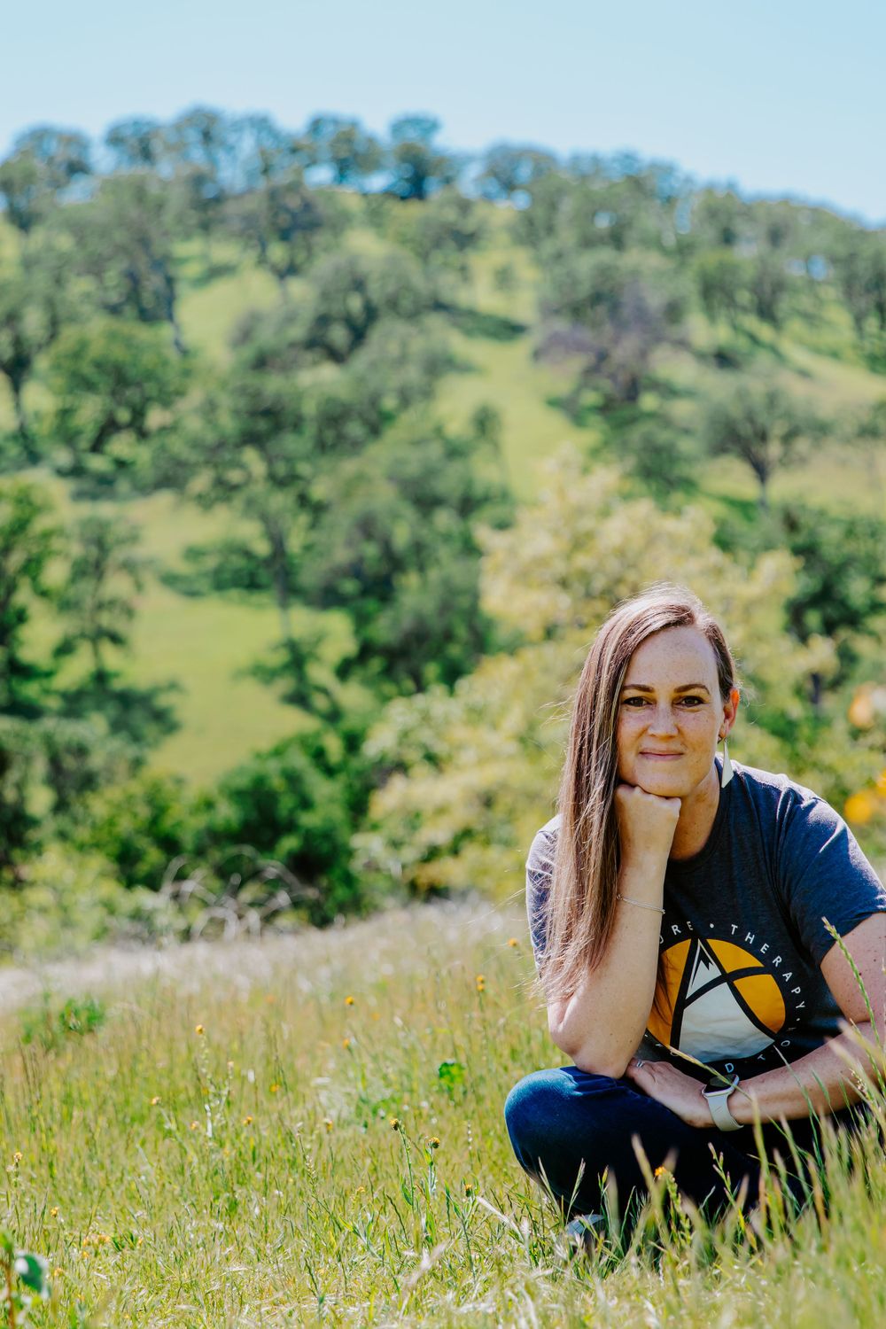 A person in a navy t-shirt sits in a sunny meadow with rolling green hills in the background.