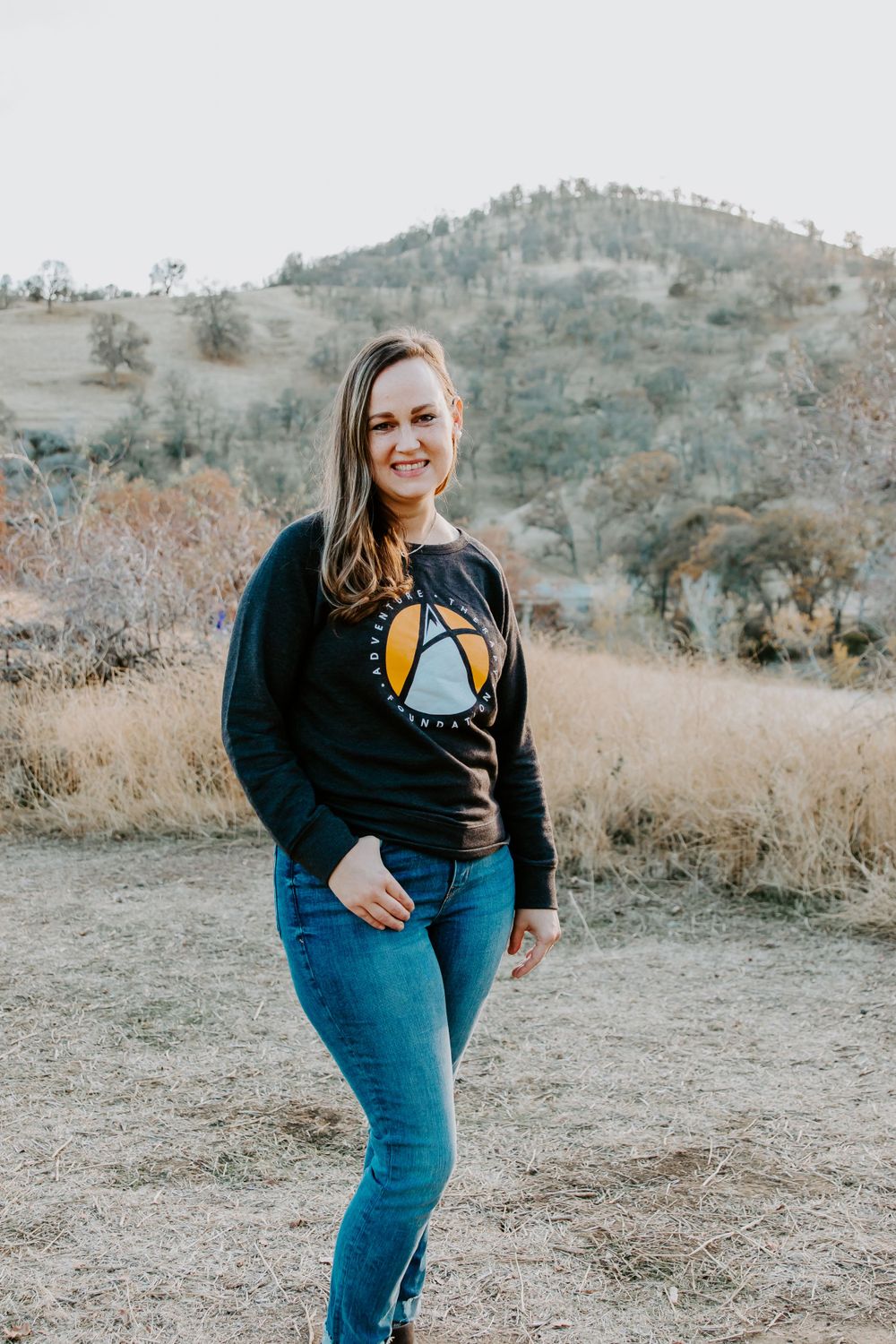 Person in dark sweatshirt and jeans posing on hiking trail with rolling hills in background.