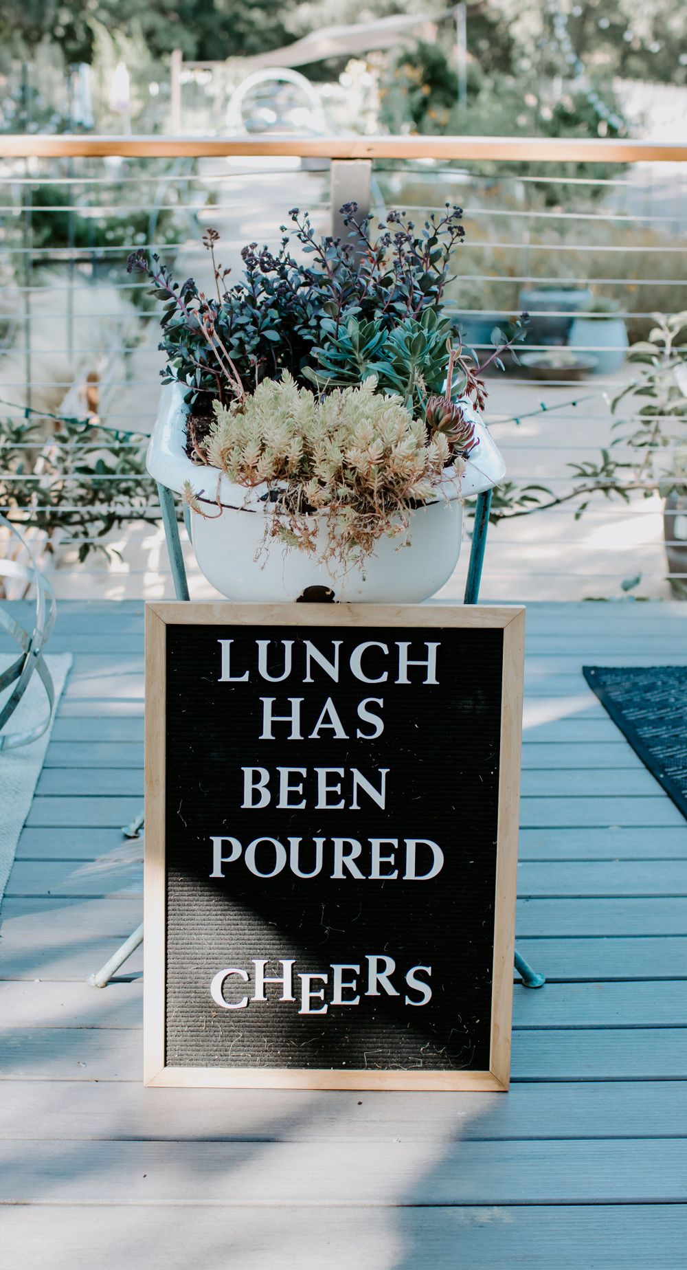 A letterboard sign reading 'Lunch has been poured, cheers' with floral arrangement on outdoor deck.