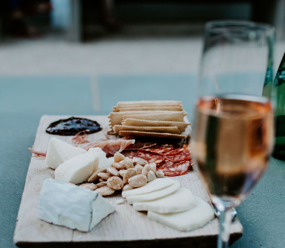 A cheese and charcuterie board with crackers, meats, and a glass of rose wine on a gray surface.