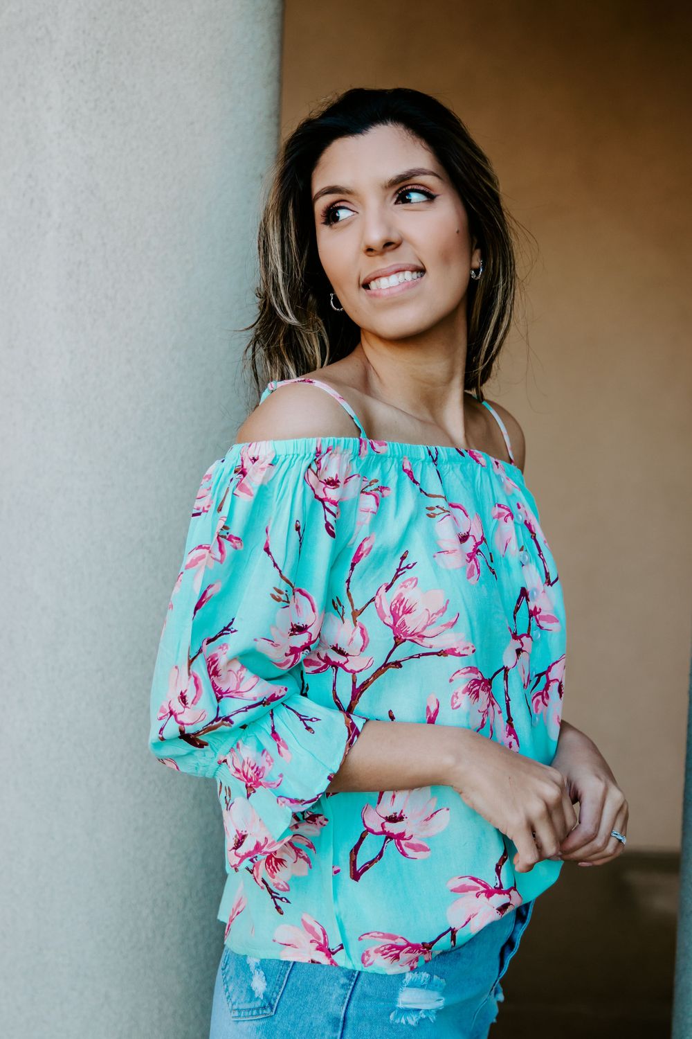 Woman in turquoise floral off-shoulder top and distressed jeans poses against a neutral wall.