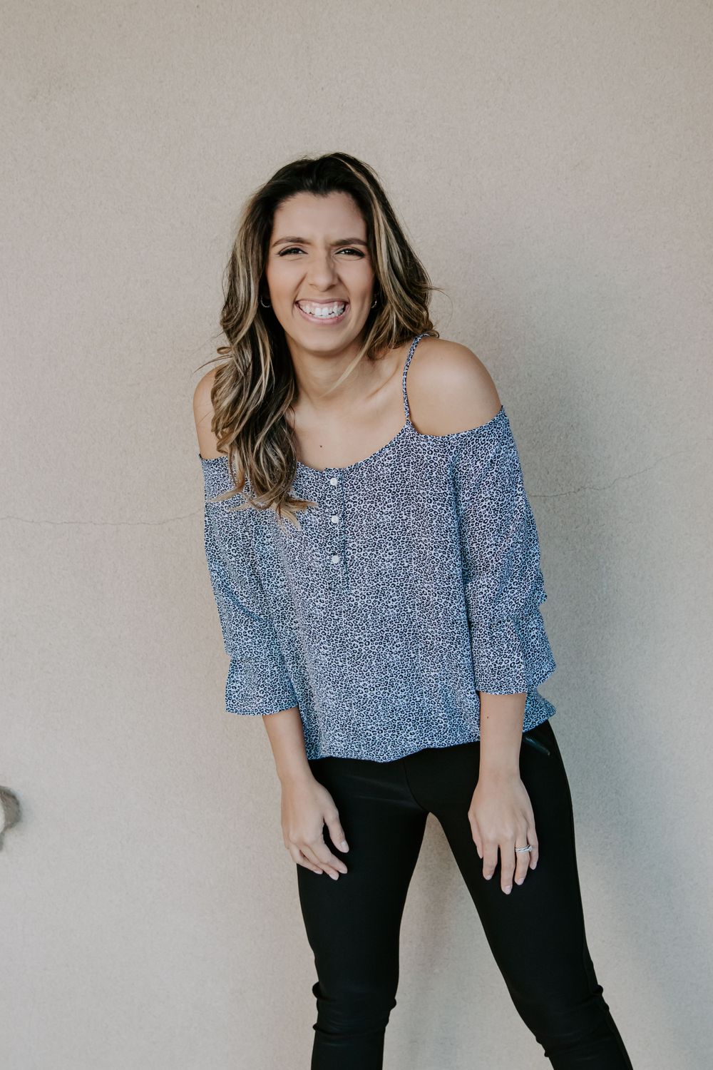 Woman wearing blue printed off-shoulder top and dark pants smiles at camera.