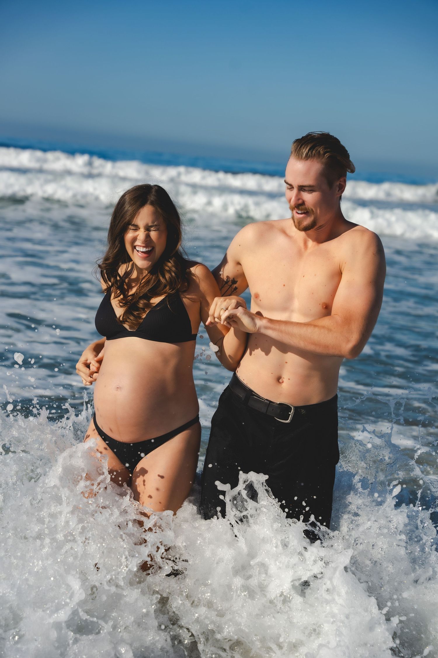 A couple laughs and splashes together in the ocean waves on a sunny summer day at the beach for their maternity photos.