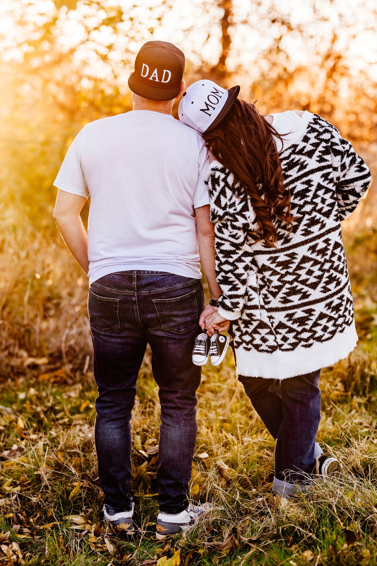 Couple wearing Dad and Mom hats holding baby shoes at golden hour.