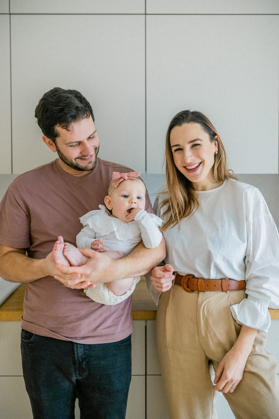 Family with infant sharing a joyful moment together against a light wall in casual attire.