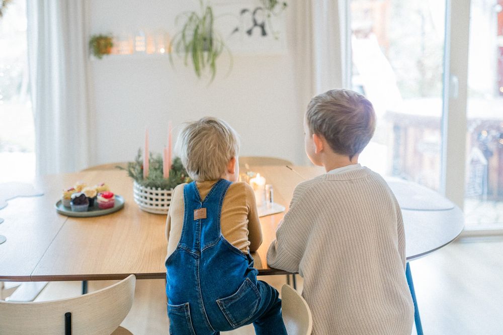Two children sitting at dining table looking out bright window with holiday decorations.
