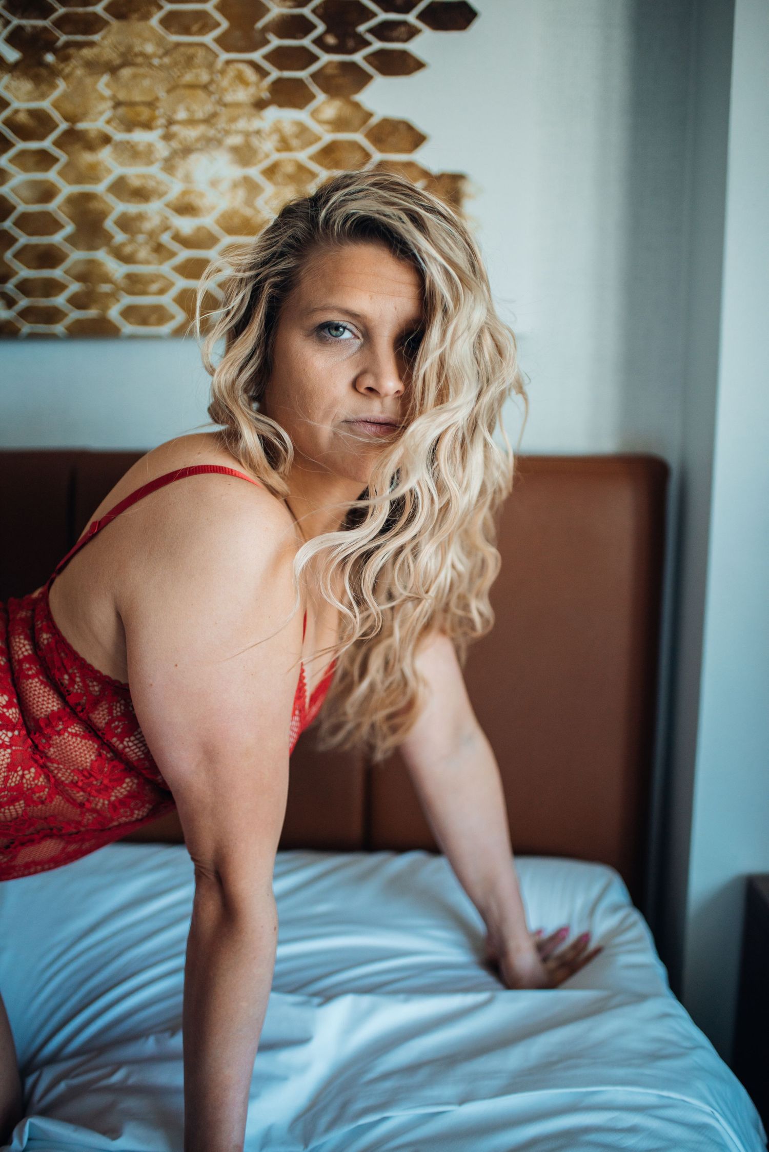 Woman in red lace poses on bed near geometric wall art.