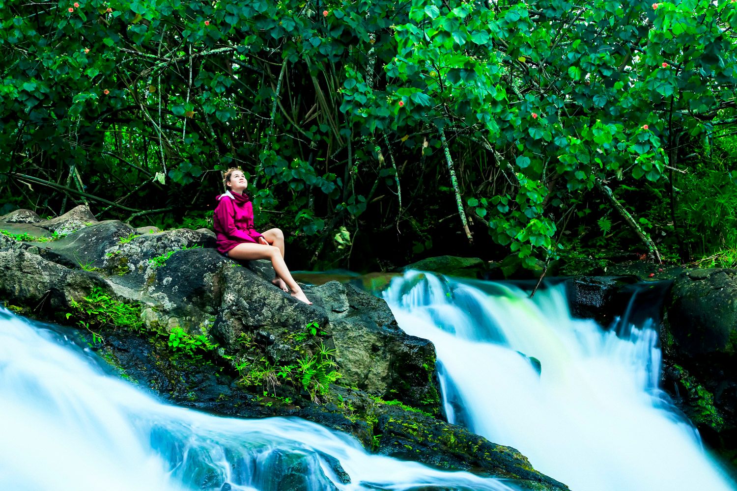 girl sitting on rocks in the middle of a waterfall in Kauai Hawaii wearing bright purple