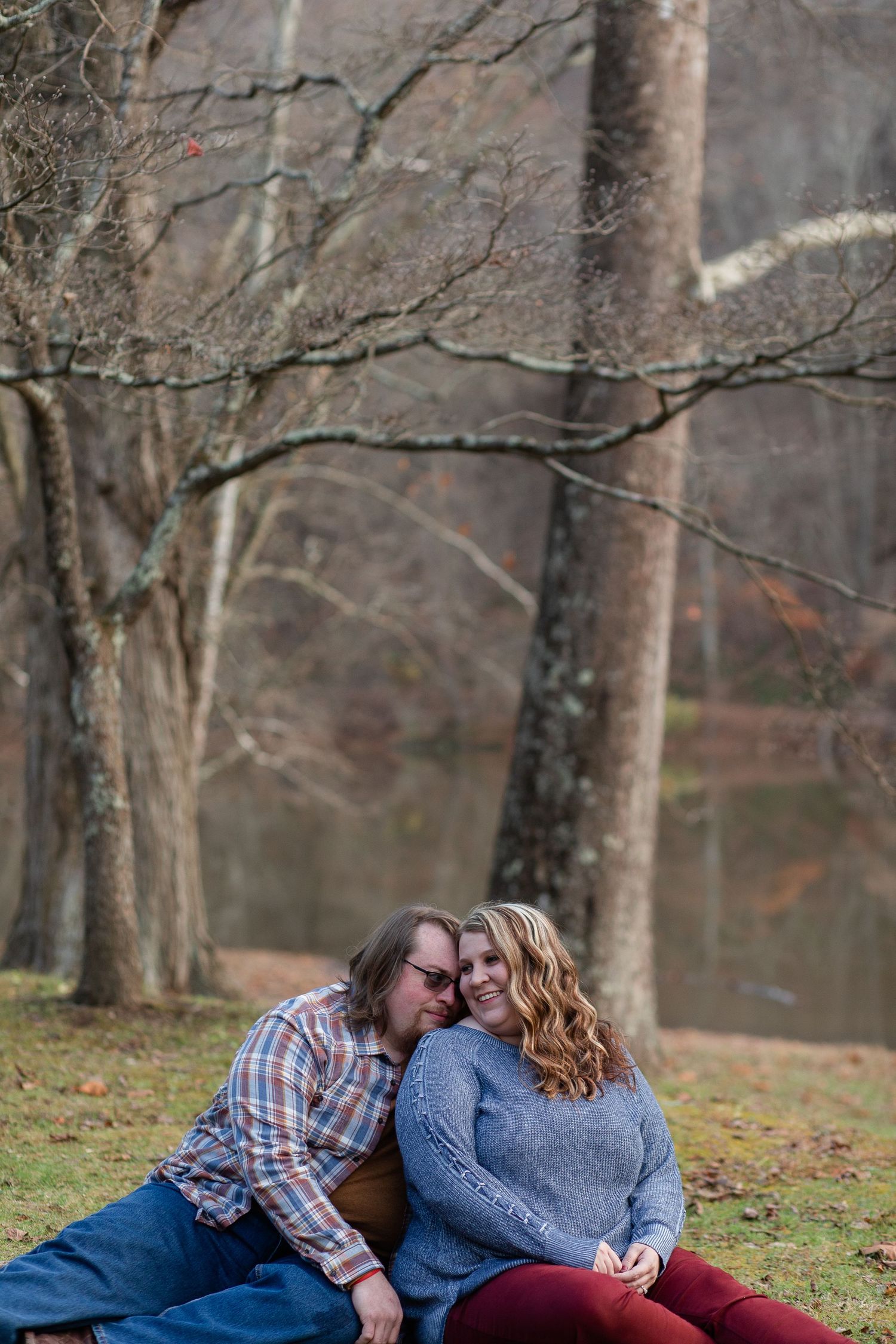 A couple lies together on grass near a lake during fall season.