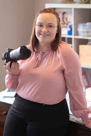 Professional photographer holding a DSLR camera wearing a pink blouse and black pants in a home office setting.