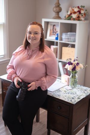 A photographer in pink blouse stands beside a wooden desk with camera equipment and floral decor.
