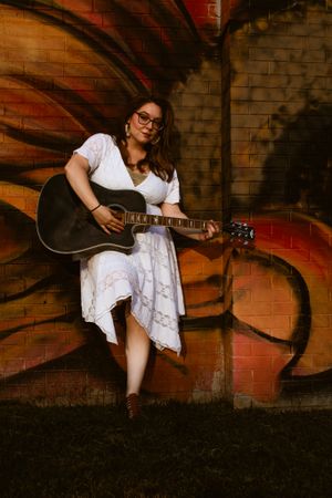 Musician in white lace dress poses with guitar against urban brick wall with orange graffiti.