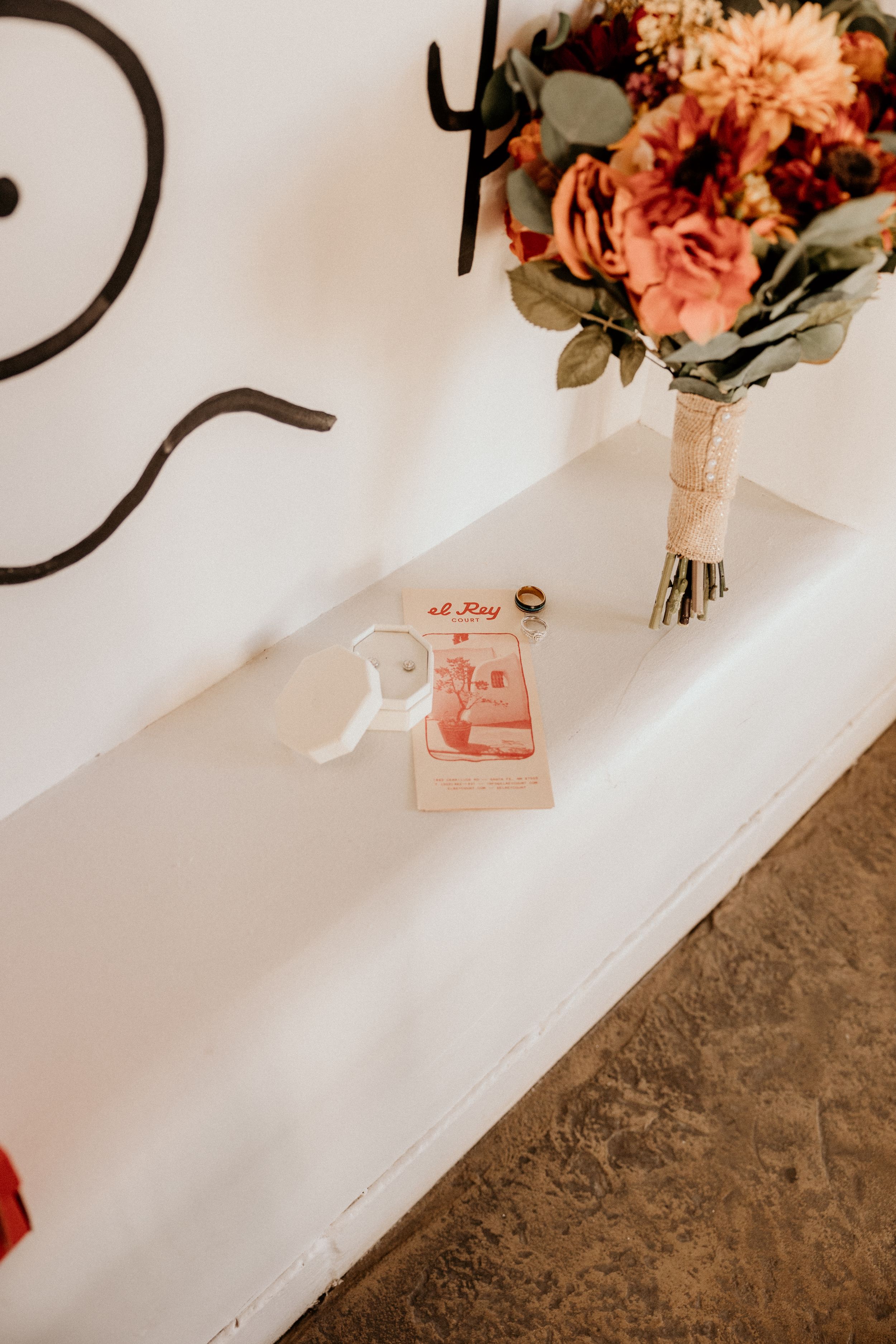 Wedding flat lay with autumn floral bouquet, rings, and El Rey Court brochure on white surface.