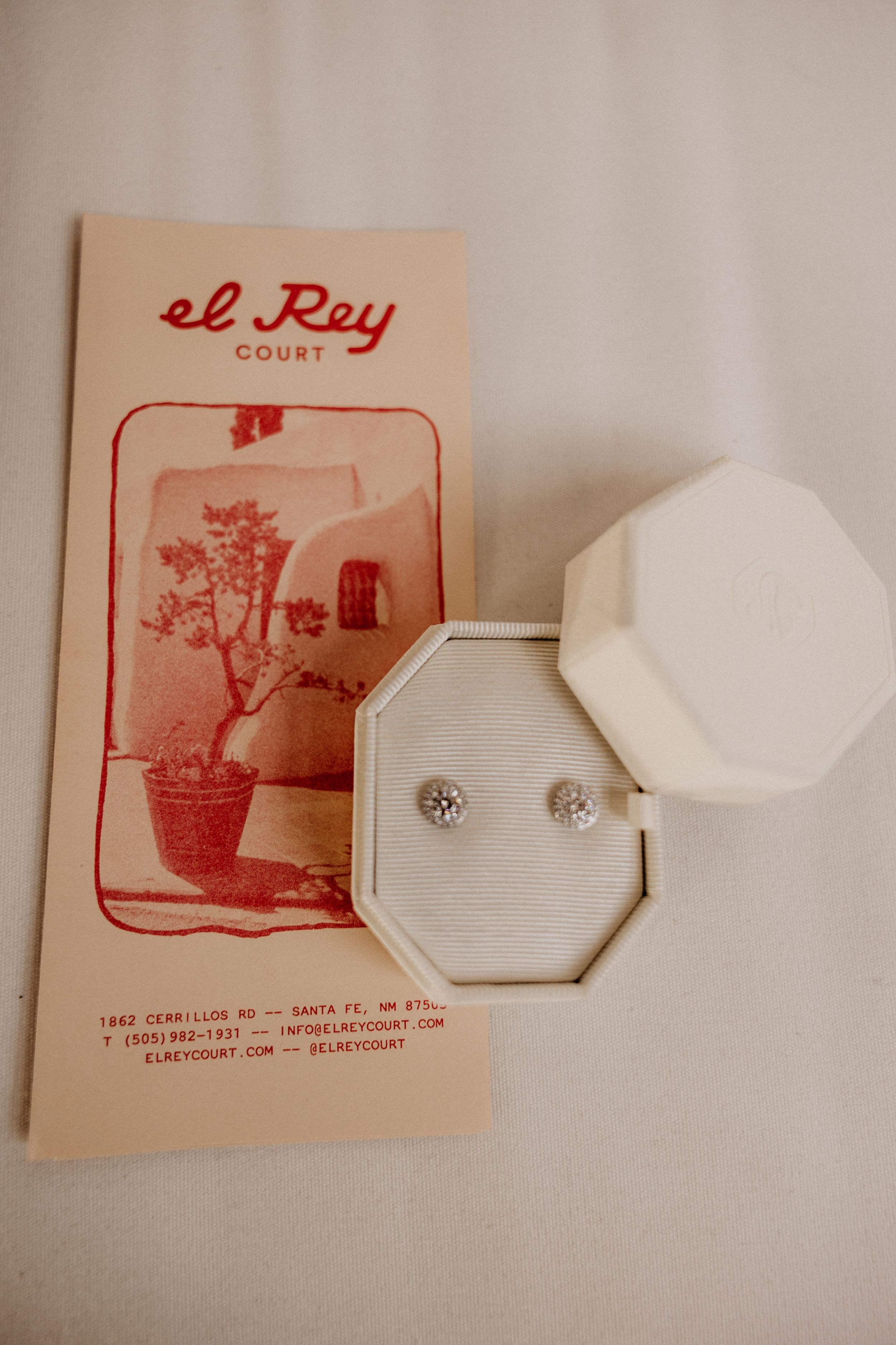 Diamond stud earrings in white octagonal ring box beside El Rey Court Court brochure.