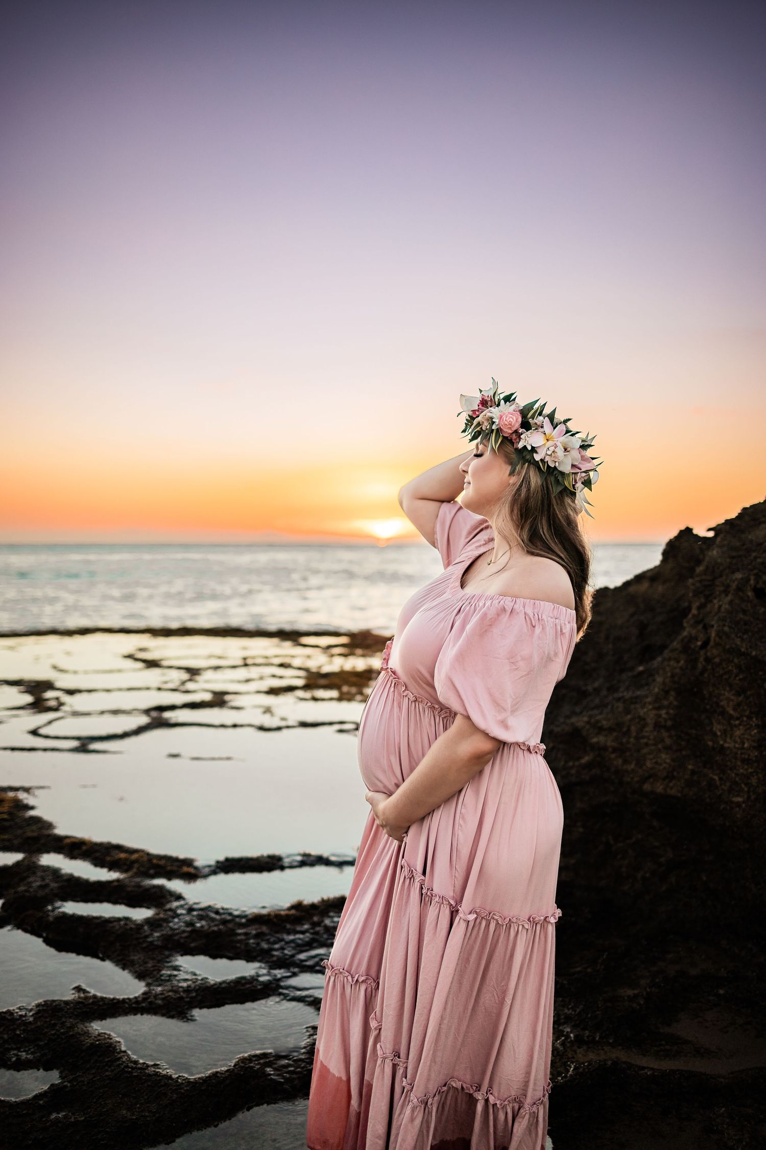 Sunset ocean view with figure in pink dress wearing floral crown against rocky coastline.