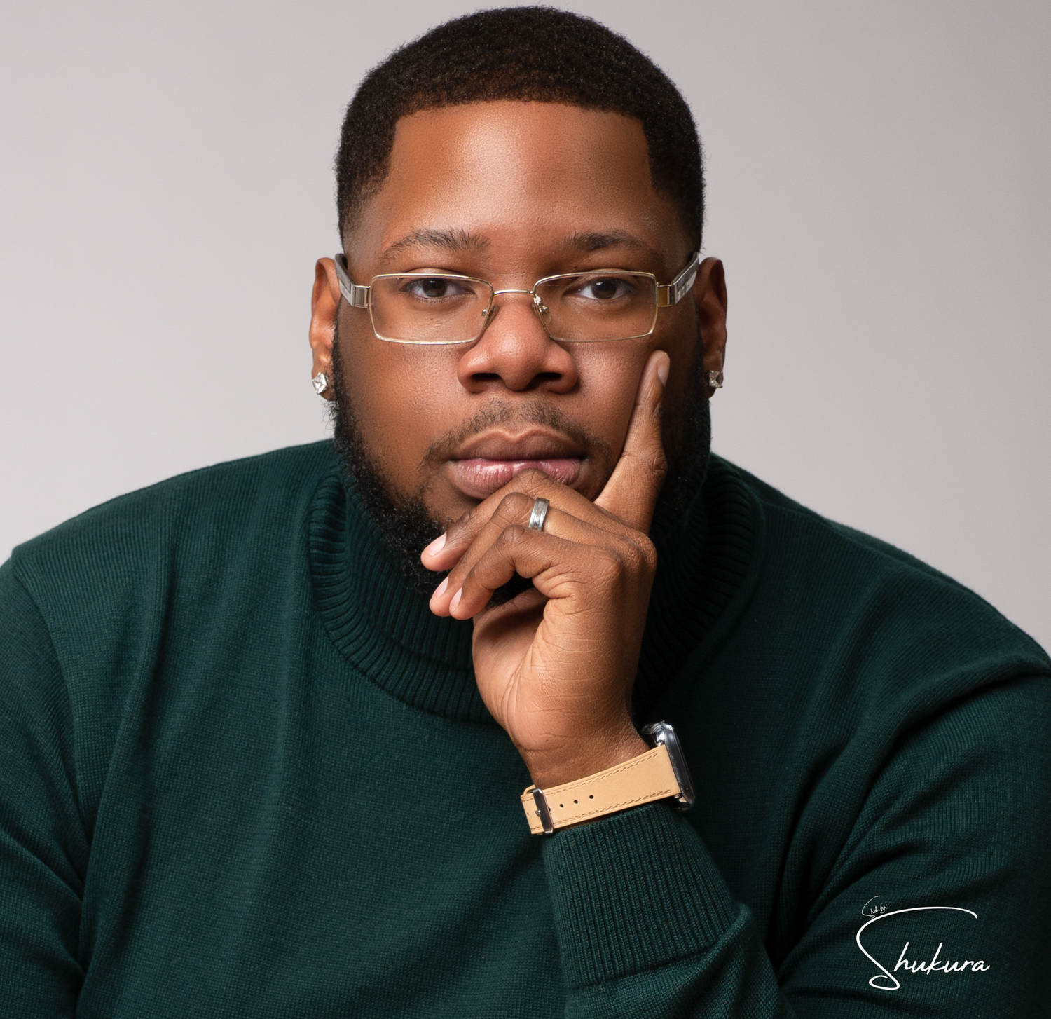 Professional portrait of person wearing glasses and dark green sweater with contemplative pose against neutral background.