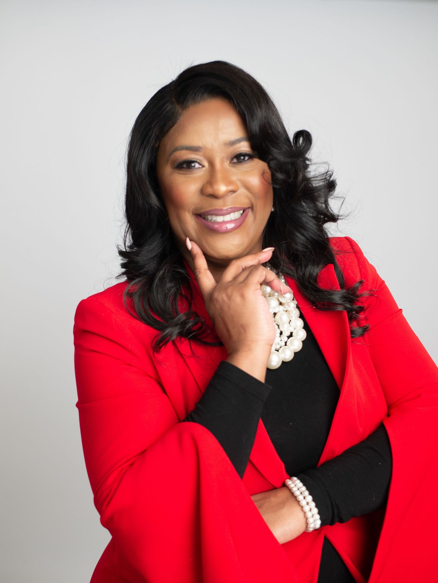 Professional portrait with person in red blazer and pearl accessories against gray background.