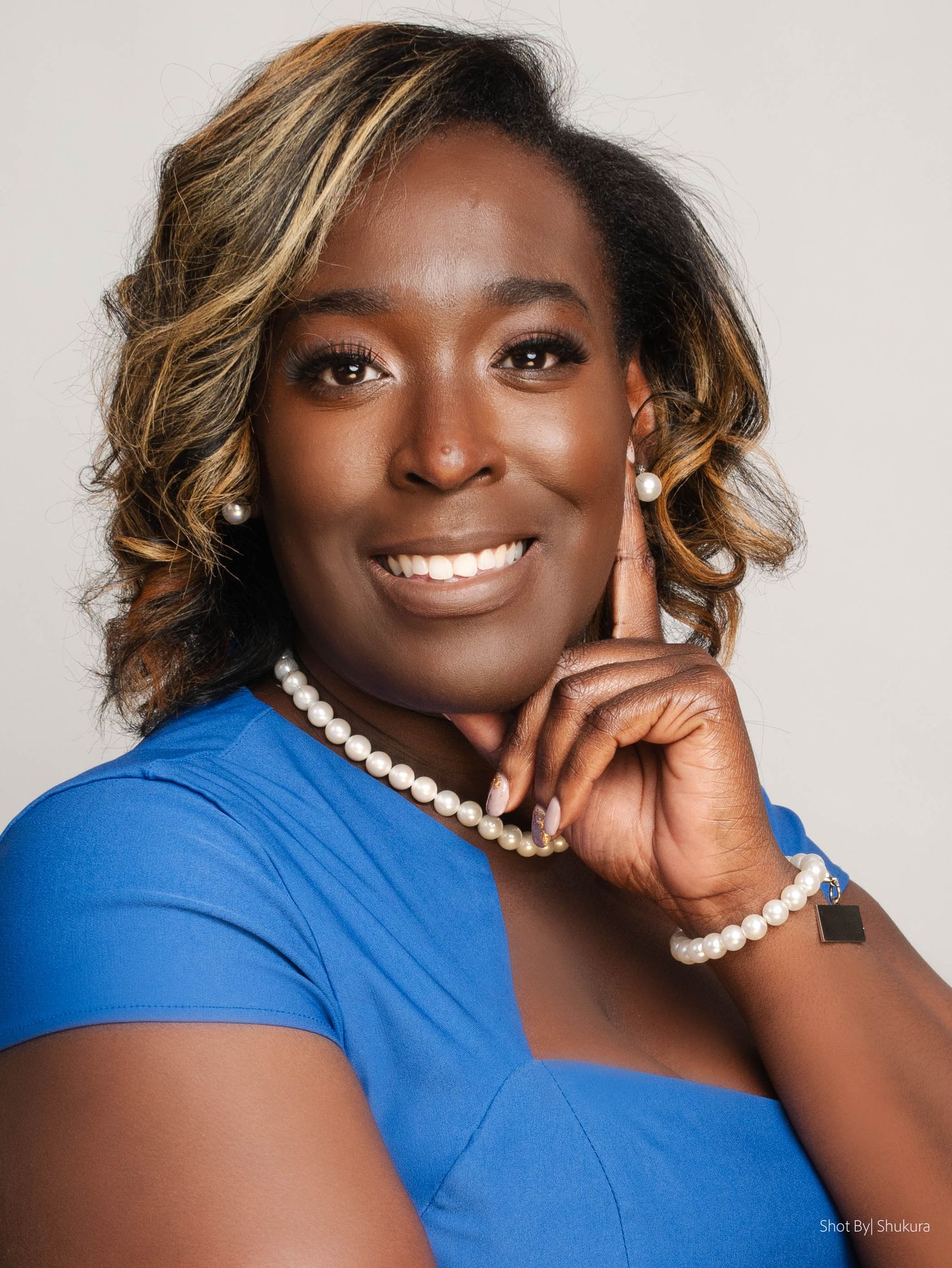 Professional headshot of someone in blue top wearing pearl jewelry against light background.