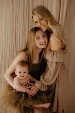 A tender studio portrait of a parent and baby in muted tones against a beige curtain backdrop.