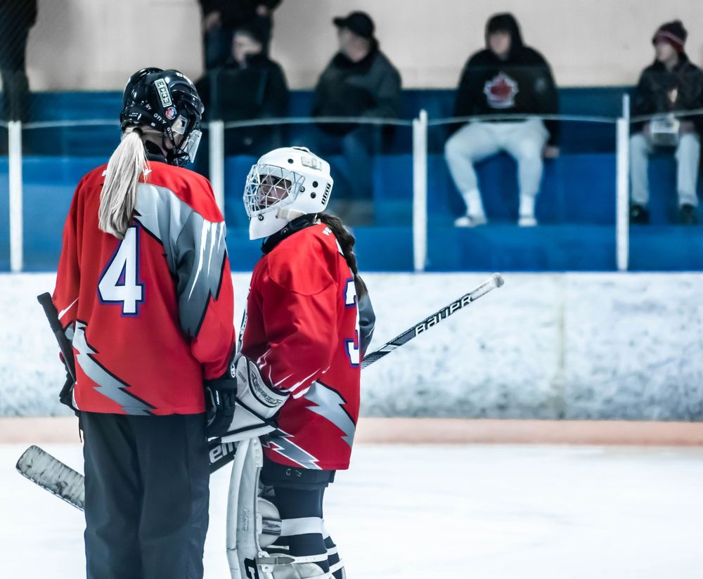 Two hockey players in red jerseys stand near the rink boards while spectators watch from above.