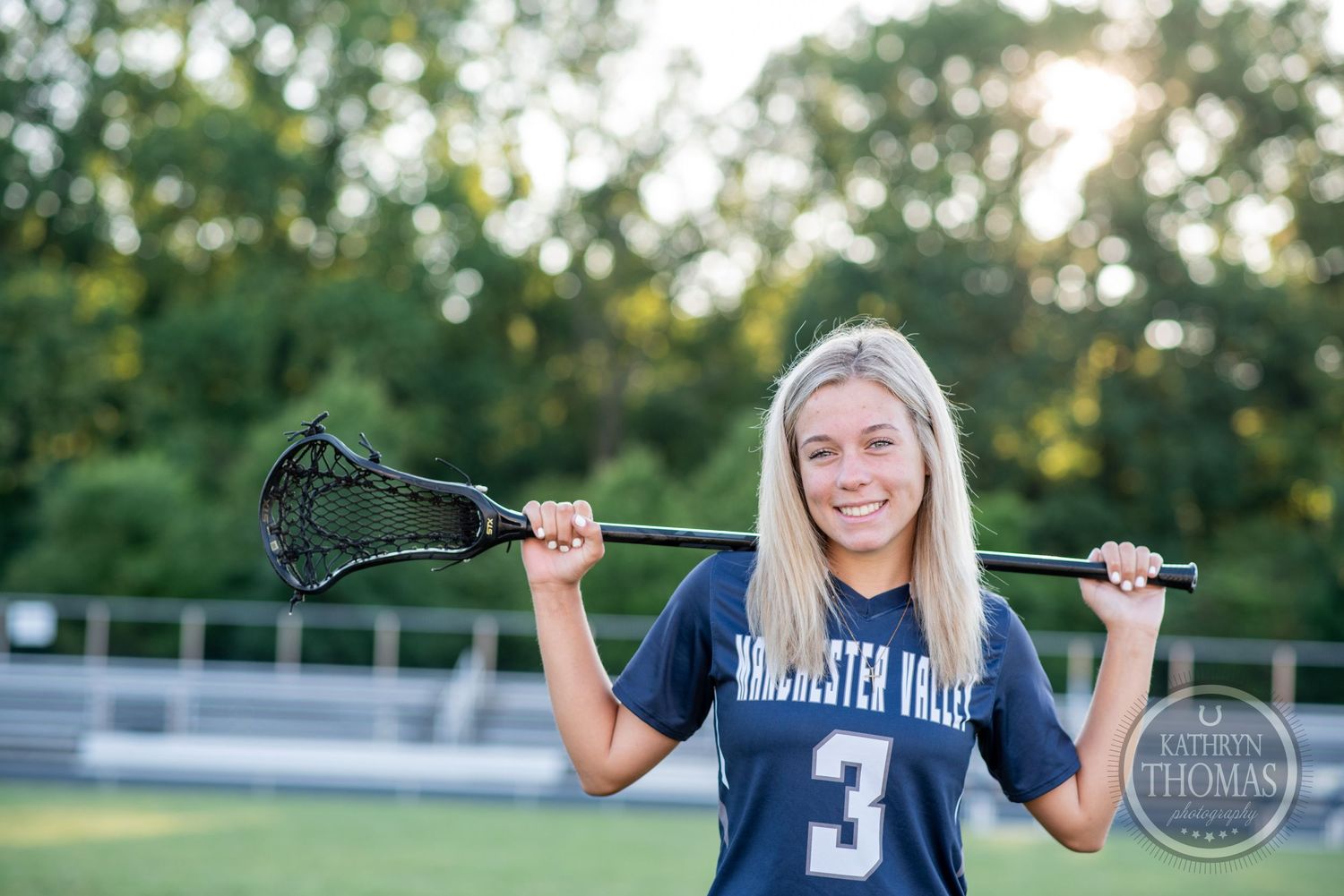 Athlete smiling while holding lacrosse stick across shoulders on sunny sports field.