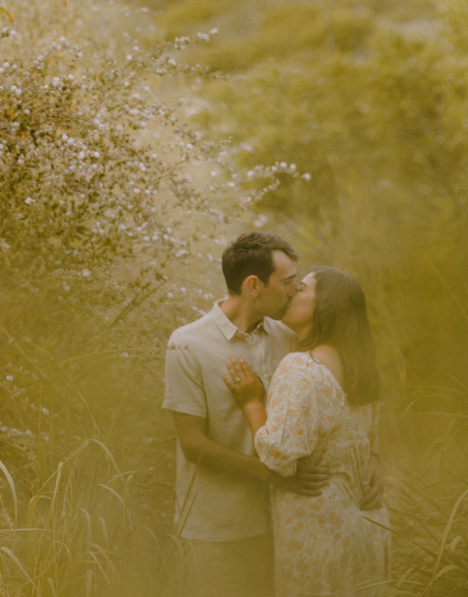 artistic shot of couple kissing under a tree with green, blurry, bokeh in the foreground