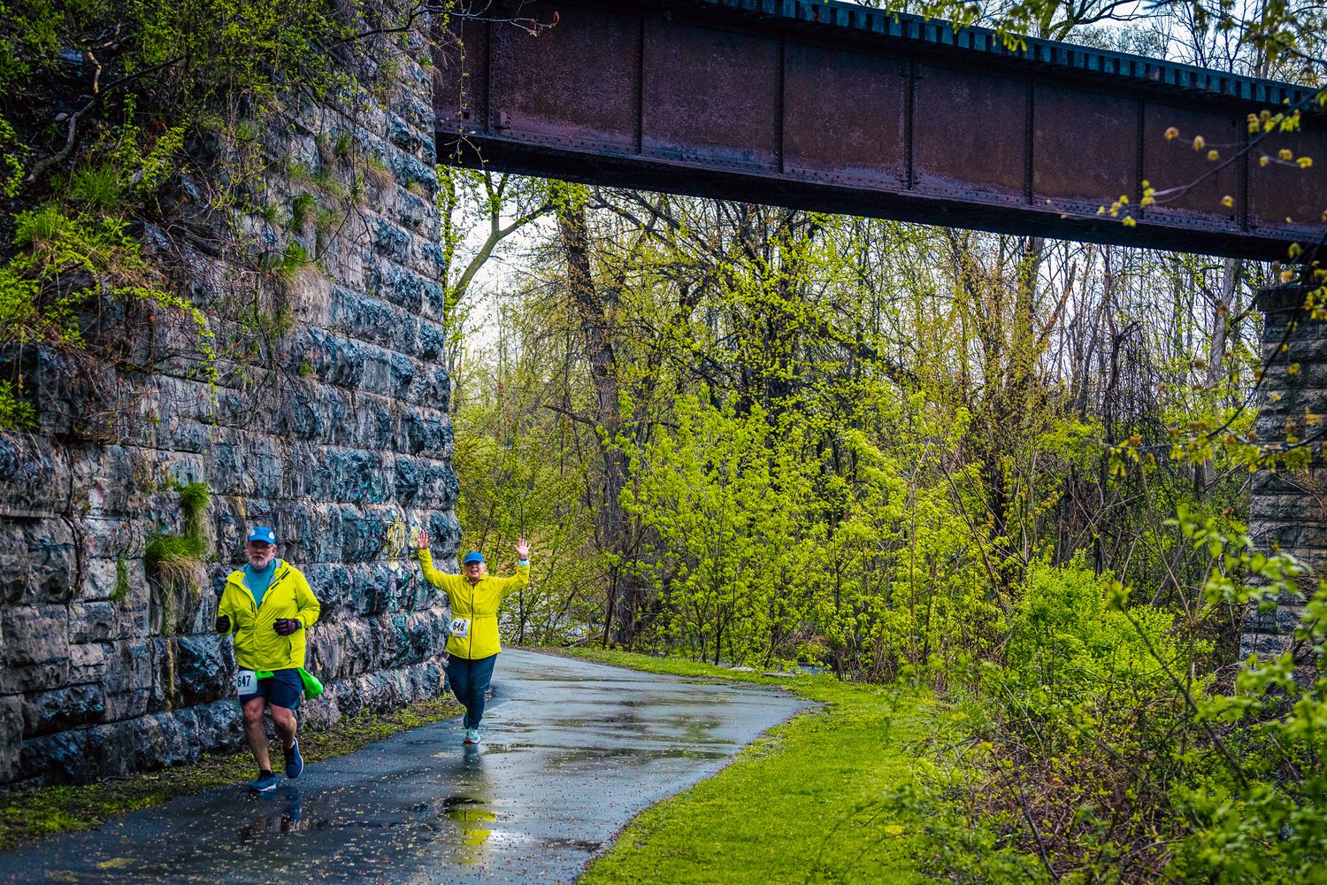 Two runners in bright jackets jogging under stone bridge on wet path.
